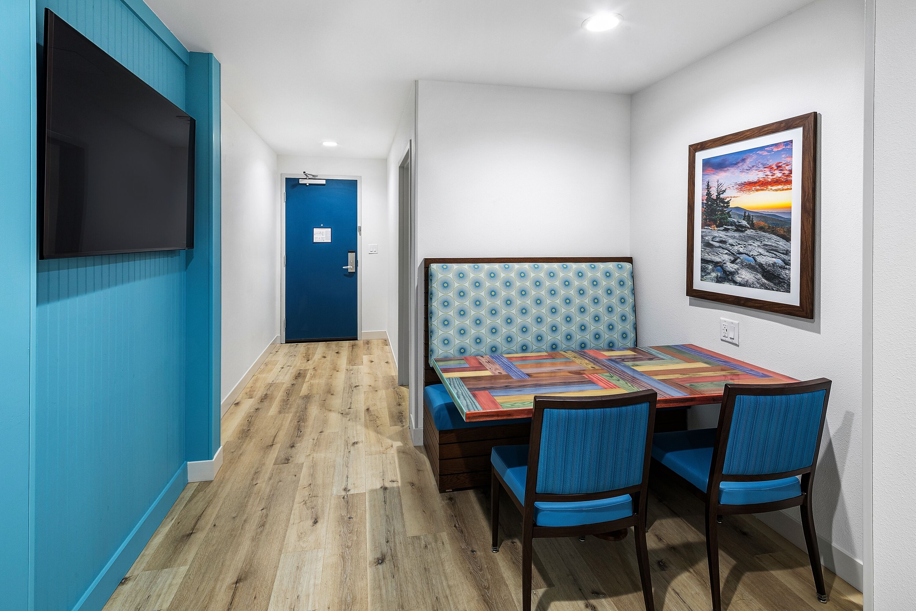 Una stanza con pavimento in legno, una porta blu, un tavolo con tovaglia colorata, due sedie azzurre e un quadro appeso al muro.