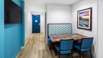 Una stanza con pavimento in legno, una porta blu, un tavolo con tovaglia colorata, due sedie azzurre e un quadro appeso al muro.