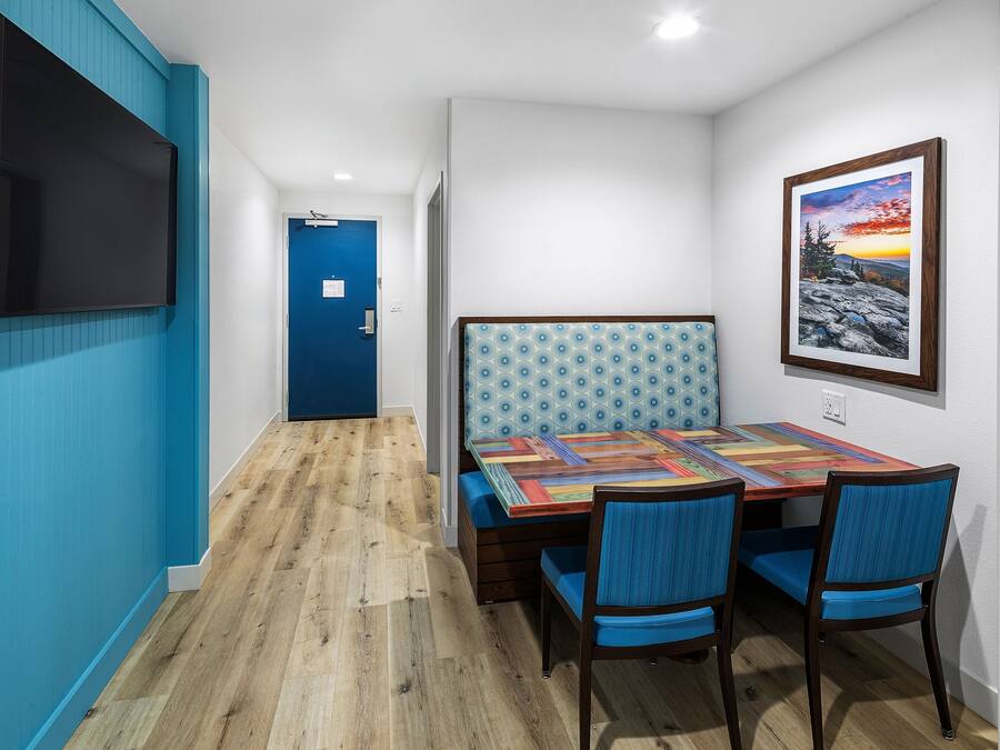 Una stanza con pavimento in legno, una porta blu, un tavolo con tovaglia colorata, due sedie azzurre e un quadro appeso al muro.