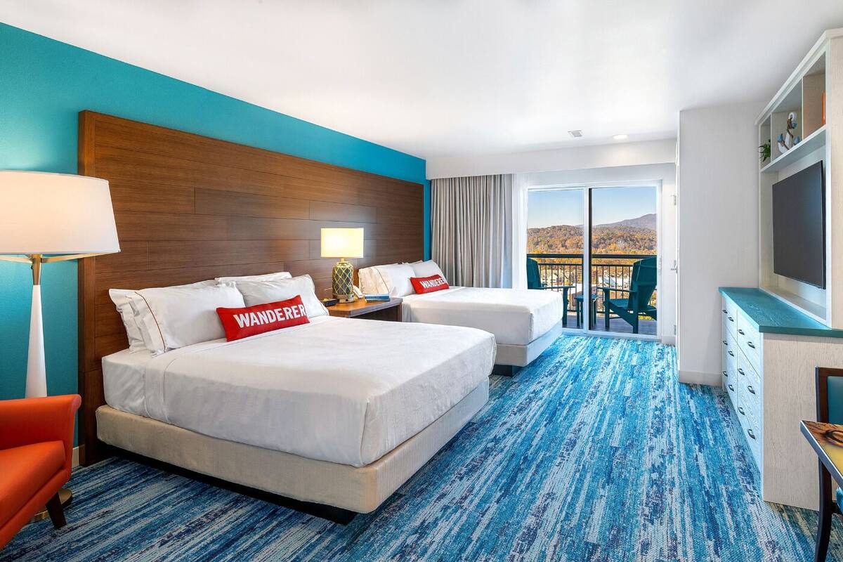 A hotel room with two beds, a TV, a desk, and a view of the outdoors.