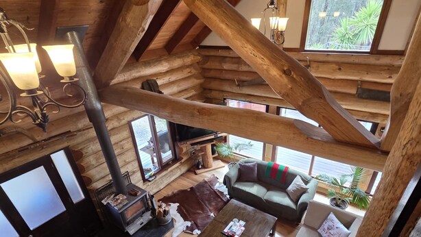 Ein geräumiges Blockhaus-Interieur mit hohen Decken, Holzbalken, einem Kamin, Ledersofas und großen Fenstern mit Blick ins Freie.