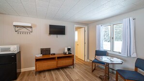 Un espace de vie compact comprenant un coin cuisine, un téléviseur à écran plat, un coin repas avec une table et des chaises, et une fenêtre avec des rideaux.