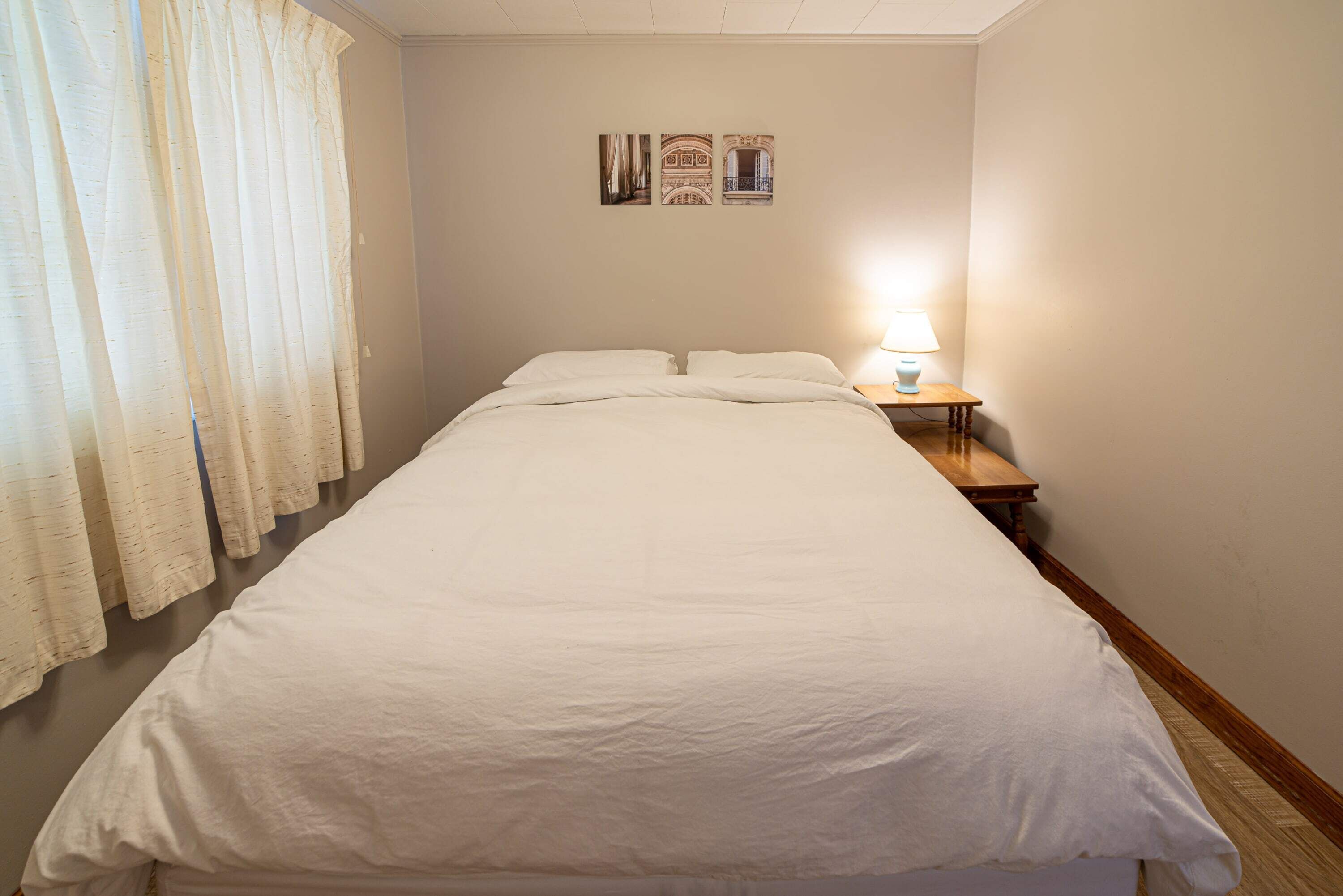 Un llit ben fet a una habitació amb una tauleta de nit, una làmpada i una finestra amb cortines.