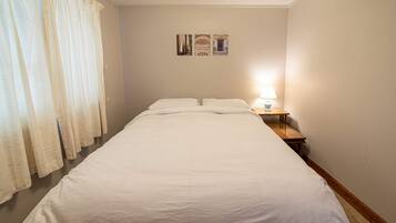 Un llit ben fet a una habitació amb una tauleta de nit, una làmpada i una finestra amb cortines.