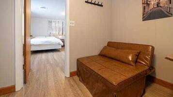 Een slaapkamer met een bruine slaapbank, een houten vloer, een bed op de achtergrond en een wandhaak.