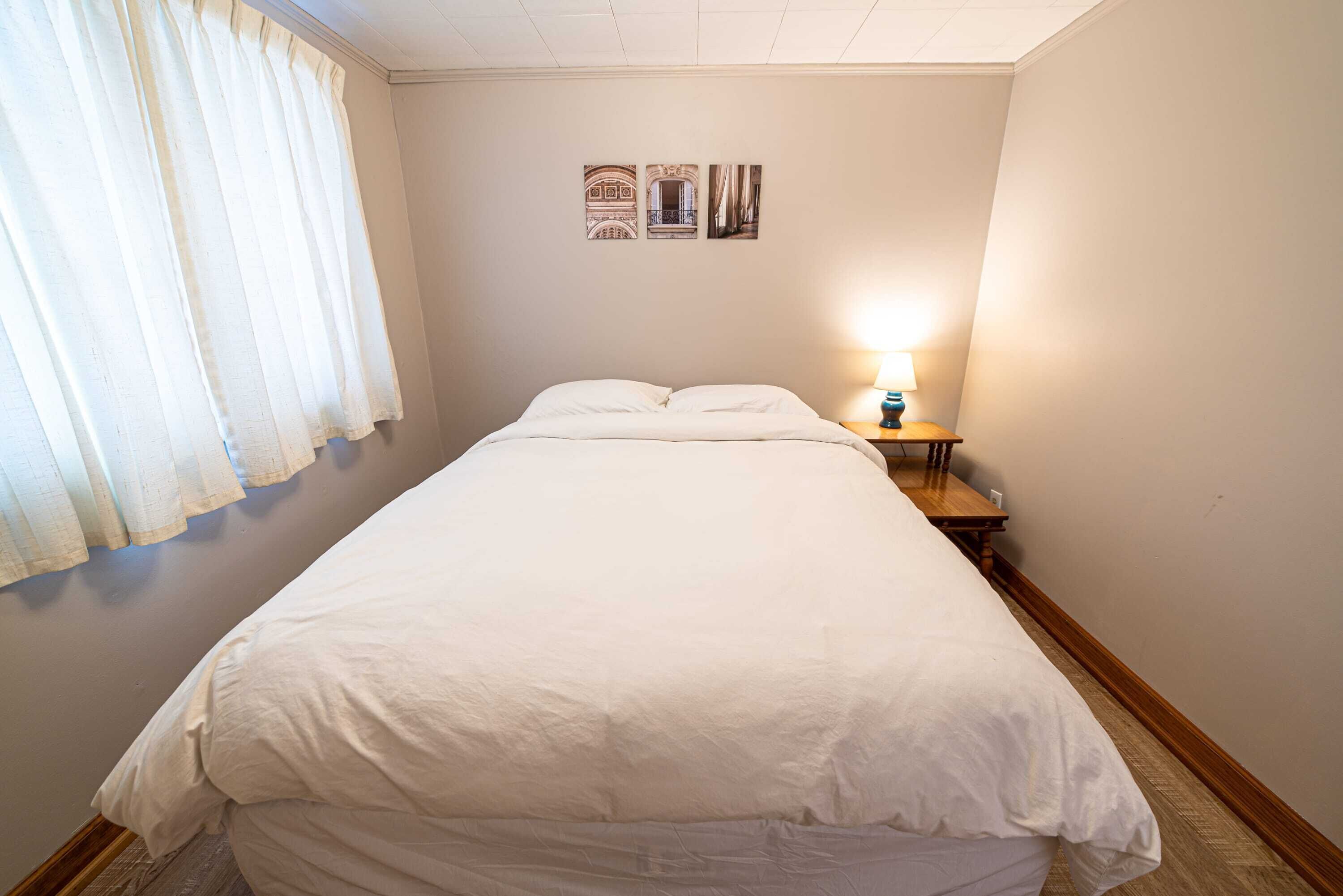 A neatly made bed in a hotel room with a nightstand, a lamp, and a window with curtains.