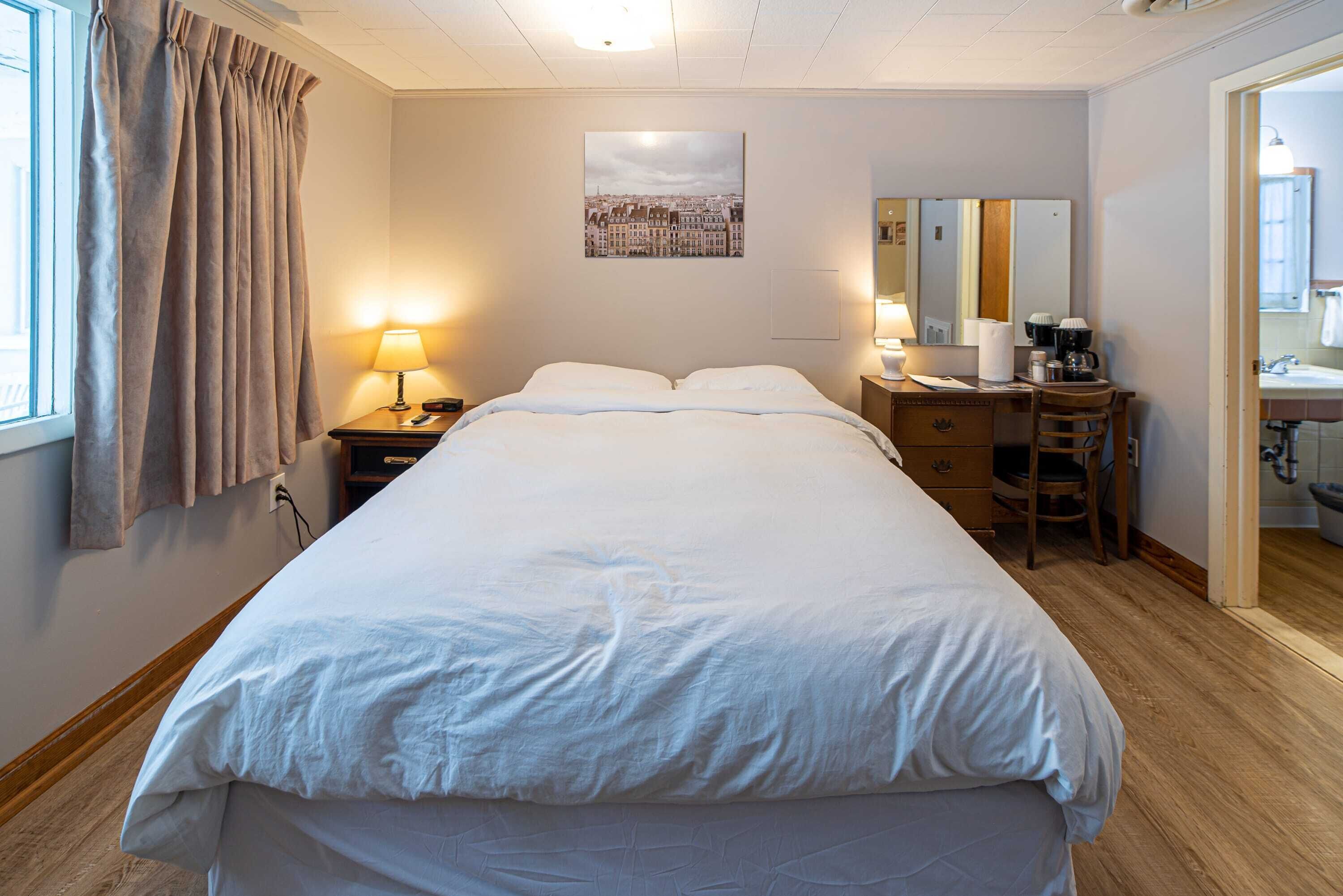 Una habitación de hotel con una cama grande, un escritorio con silla, un espejo y una ventana con cortinas.