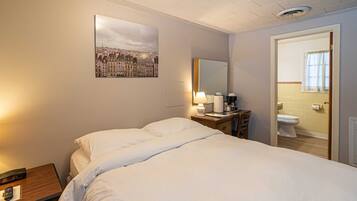 Camera d'albergo con un letto, un comodino, una scrivania con una lampada, uno specchio e un bagno visibile attraverso una porta aperta.