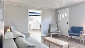 Een moderne woonkamer met een grijze bank, een houten salontafel en twee stoelen. Een keuken is zichtbaar door een open doorgang.