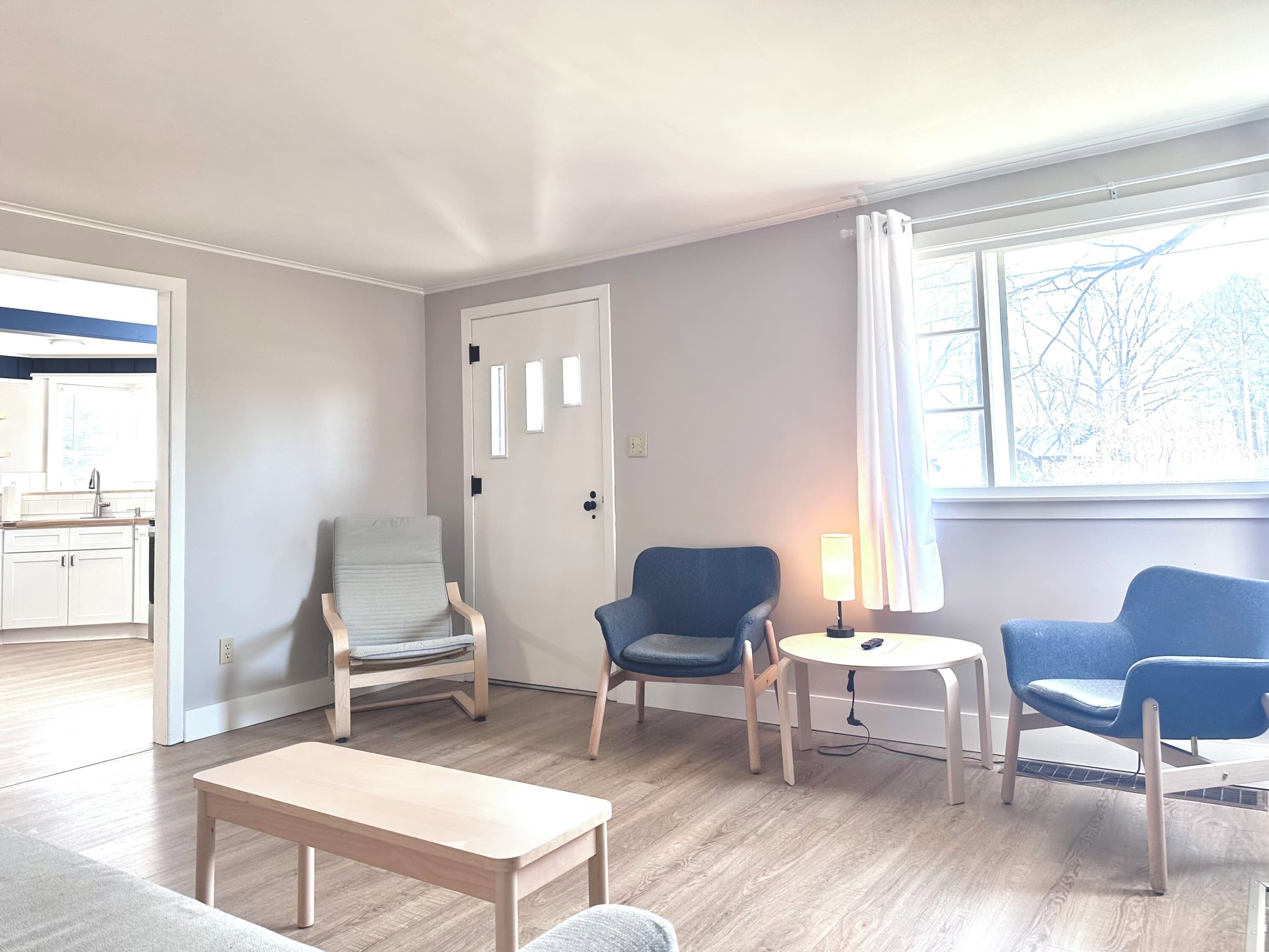 Een moderne woonkamer met een houten vloer, een klein rond tafeltje met een lamp, twee blauwe stoelen, een houten bankje en een deur die naar een keuken leidt.