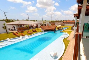 A swimming pool with lounge chairs, a thatched-roof structure, and wind turbines in the background. - POUSADA SOL E VIOLÃO (São Miguel do Gostoso)