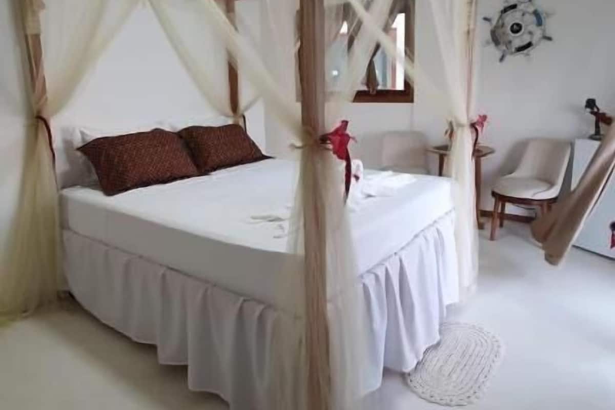 A four-poster canopy bed with white bedding and brown pillows, a small table with a chair, and a decorative wall piece.