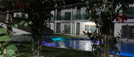 A poolside area with a mosaic-tiled deck, a thatched roof structure, and a view of a building with a pool.