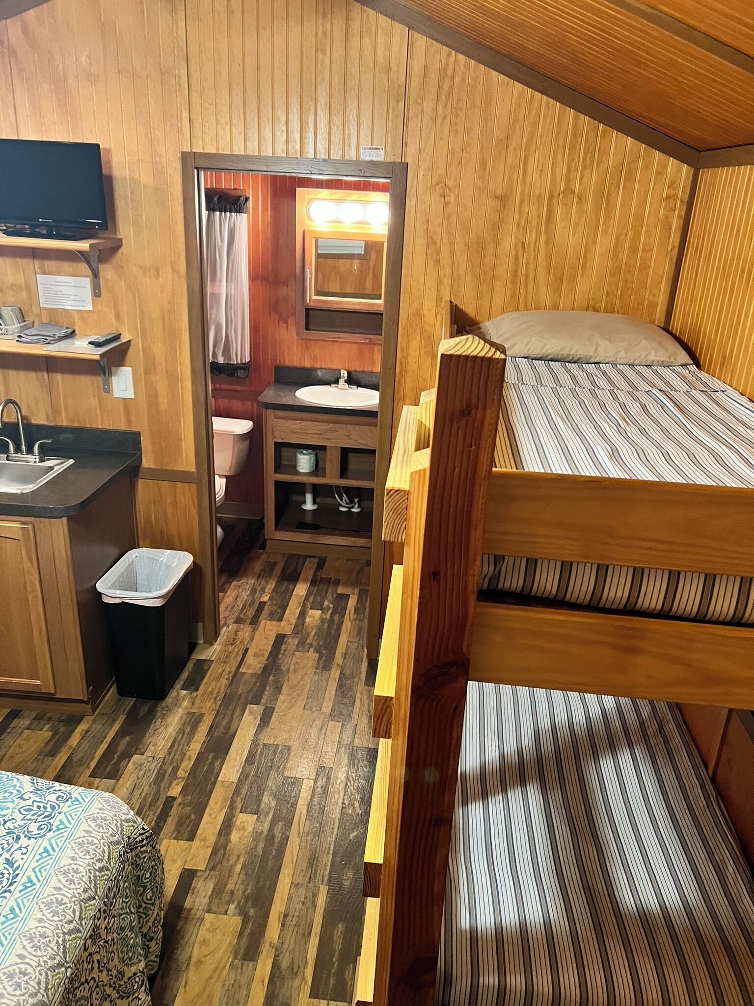 A compact, wooden-furnished room with bunk beds, a small kitchenette, a sink, and a mirror.