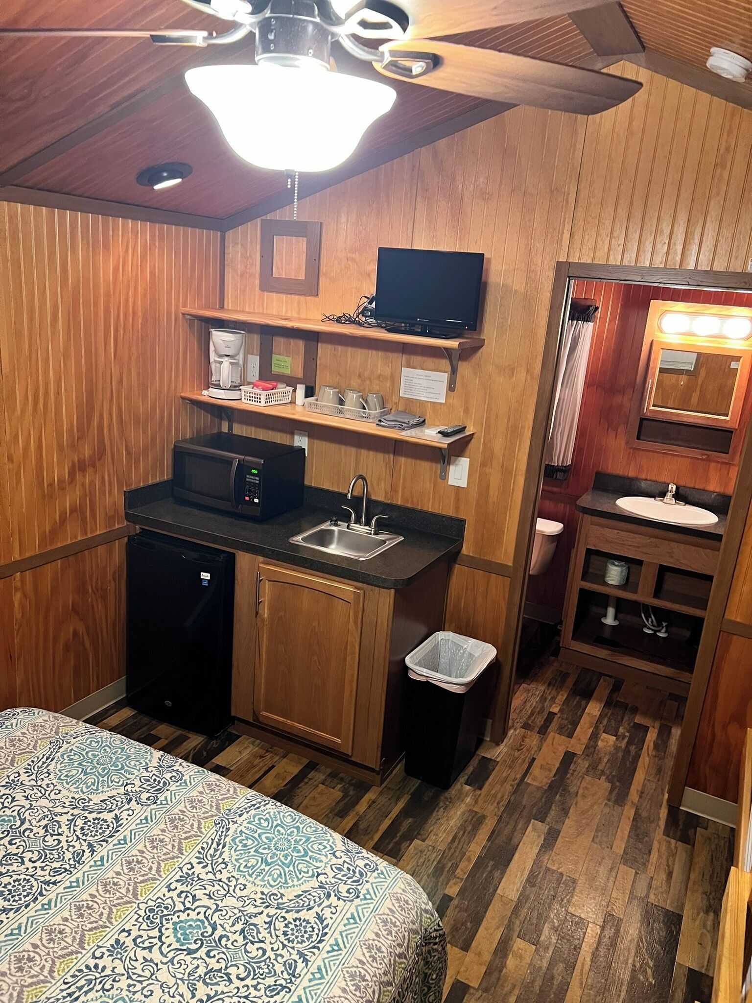 A compact kitchenette with a microwave, sink, and coffee maker, a small trash bin, and a bed with patterned bedding.