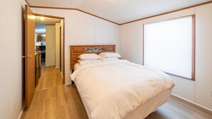 A bedroom with a wooden headboard, a large window with blinds, and a wooden floor. - Star Gate Mobile Home 16 Indian River, MI (Indian River)
