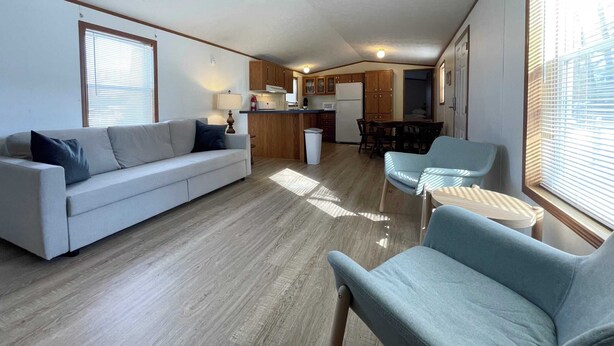 A compact living space with a sofa, two armchairs, a dining table, and a kitchenette. - Star Gate Mobile Home 16 Indian River, MI (Indian River)