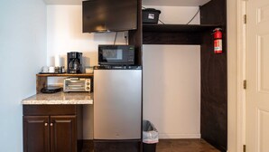 Una kitchenette compacta con microondas, tostadora y cafetera sobre una encimera de madera. Un refrigerador está ubicado contra la pared, y se observa un cubo de basura.