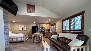 Een compacte woonruimte met een bed, een eettafel met stoelen, een bank, een klein houten bankje, een keuken met houten kasten en apparatuur, en een groot raam met zonwering.