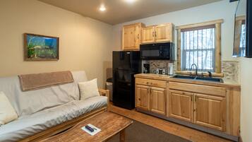 A compact living space with a sofa, a wooden table, a kitchen with wooden cabinets and appliances, and a window with a view.