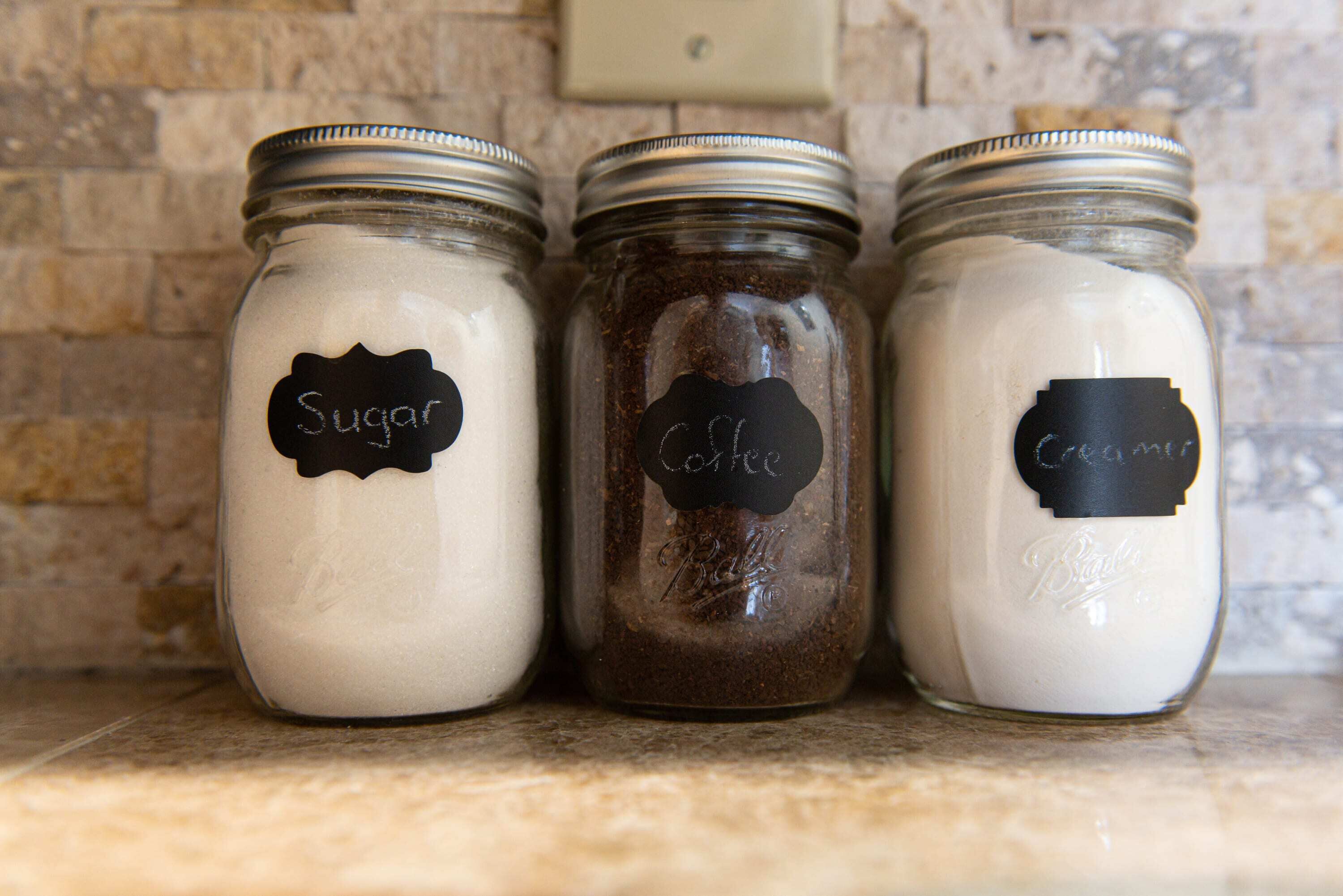 Three glass jars with metal lids, each labeled with a different type of sugar on a kitchen counter.