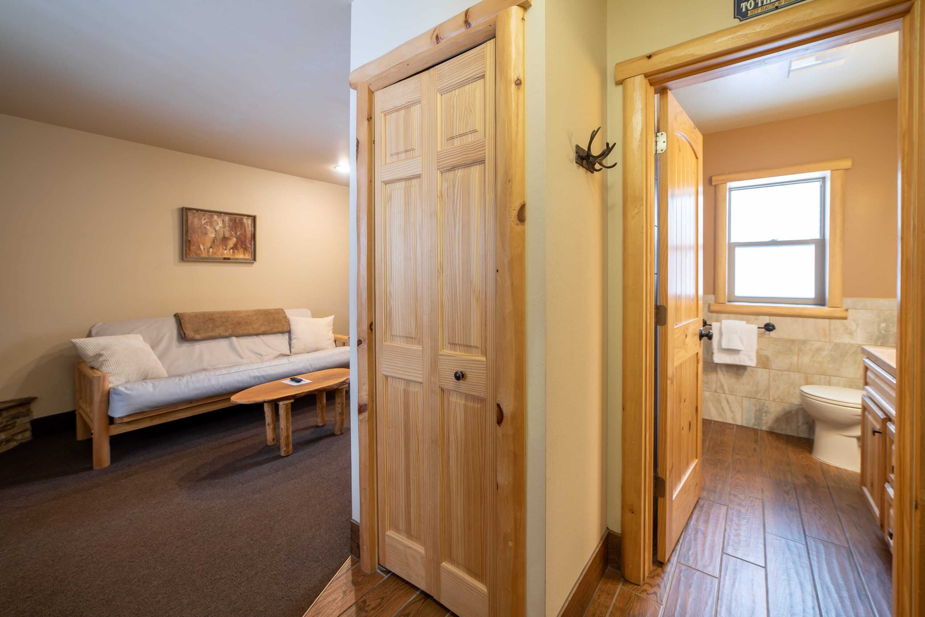 A wooden door leading to a bathroom with a toilet and a window. A wooden bench and a sofa are visible in the adjacent room.