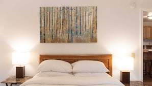 A hotel room with a wooden headboard, two bedside lamps, a large abstract painting, and a view into the kitchen area. - House at Inland Lakes Motel (Indian River)