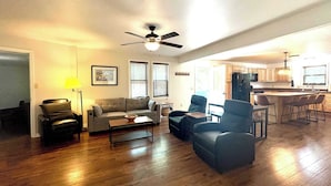 A spacious living room with a sofa, armchairs, and a coffee table. A ceiling fan and a kitchen area with a refrigerator and bar stools are visible. - House at Inland Lakes Motel (Indian River)