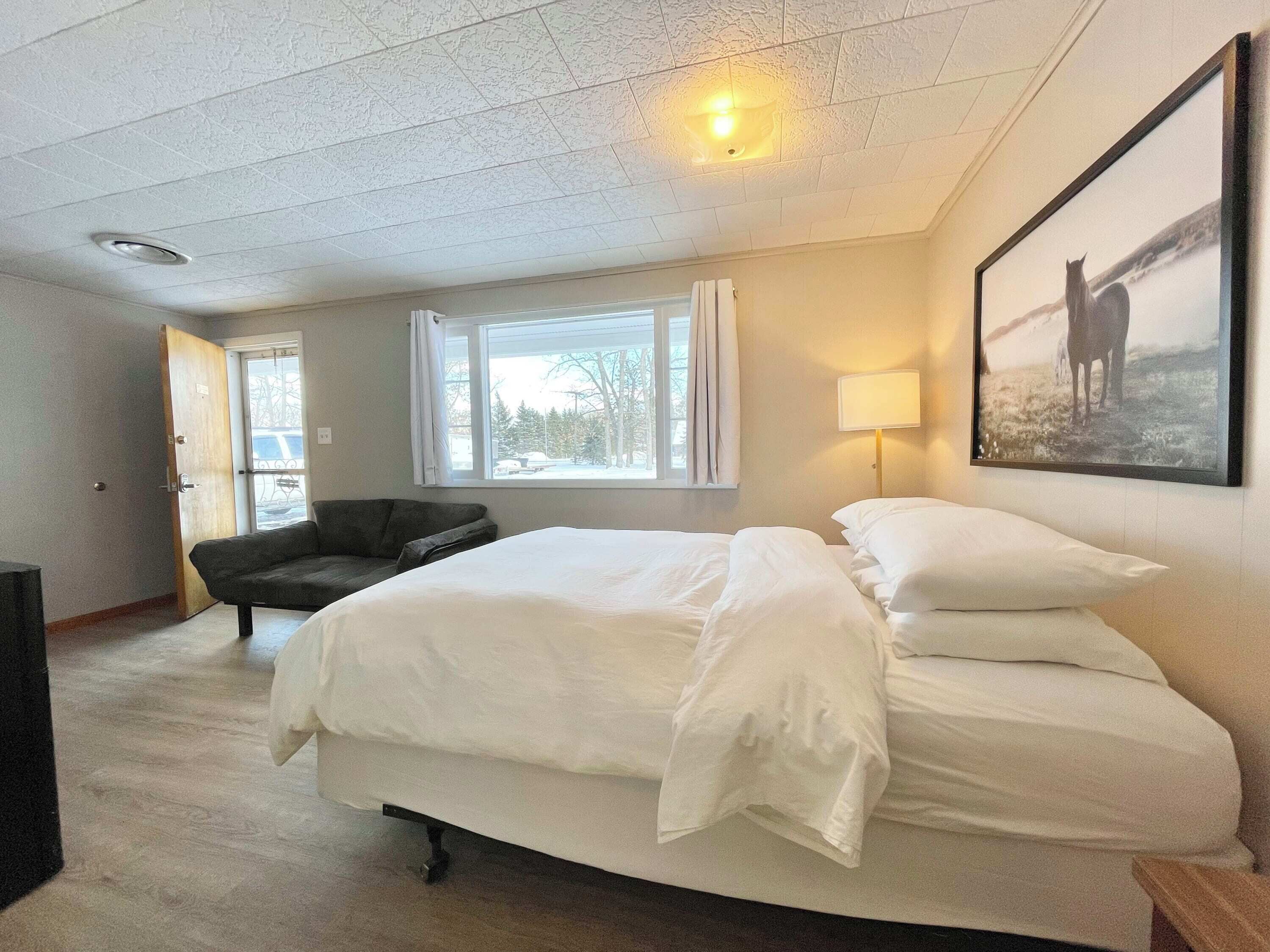 Una camera d'albergo con un letto, una sedia, una finestra con tende, una lampada e un quadro appeso al muro.