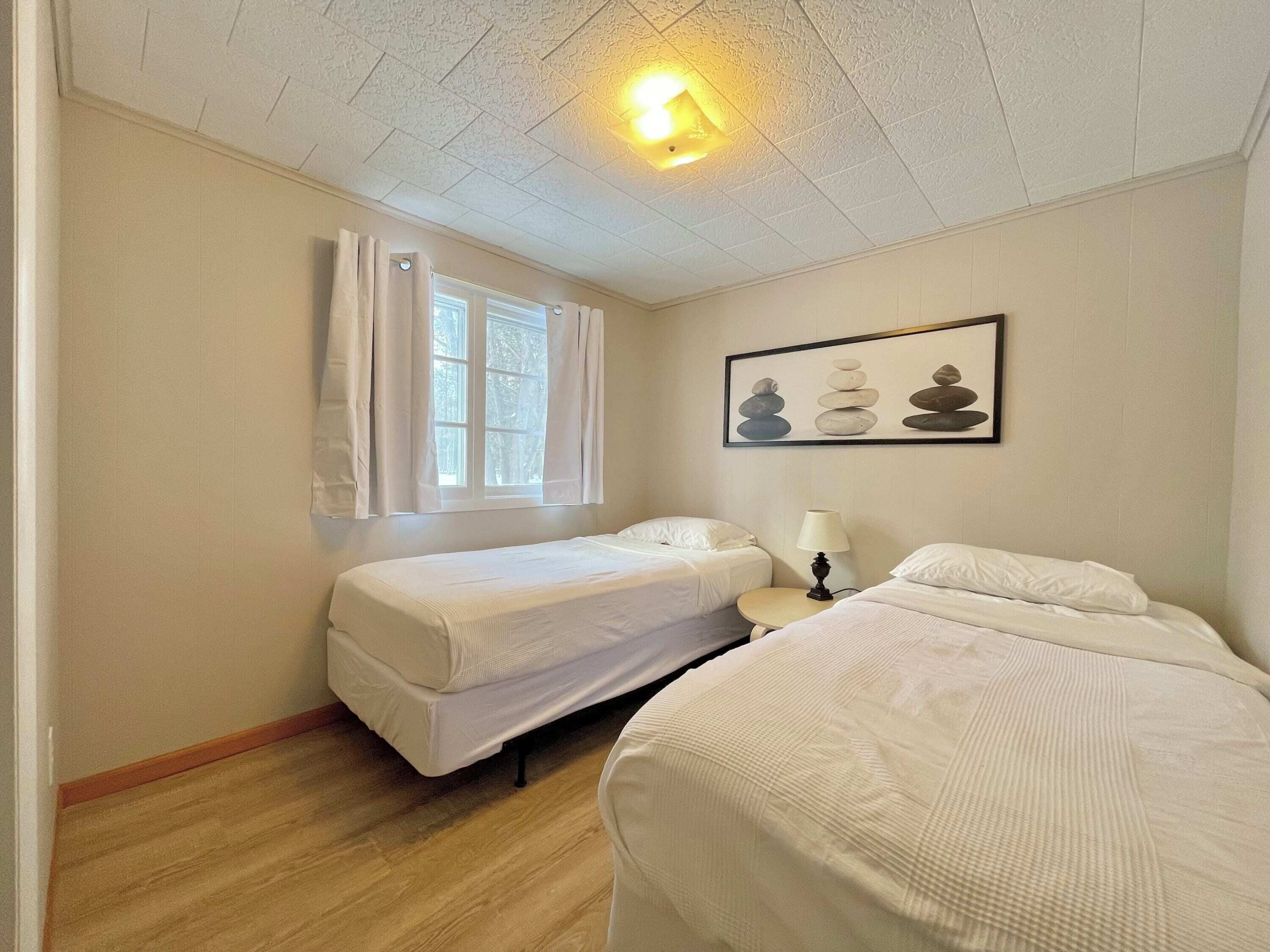 A room with two beds, a window with curtains, a ceiling light, and a framed picture on the wall.