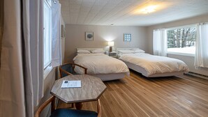 Habitación de hotel con dos camas, una mesa de madera con una silla, una mesita de noche con lámpara y una ventana con cortinas.