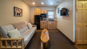 A compact living space with a sofa, a wooden coffee table, a kitchen area with wooden cabinets and appliances, and a television mounted on the wall. - The Wolverine Lodge at The Totem Lodges (Indian River)