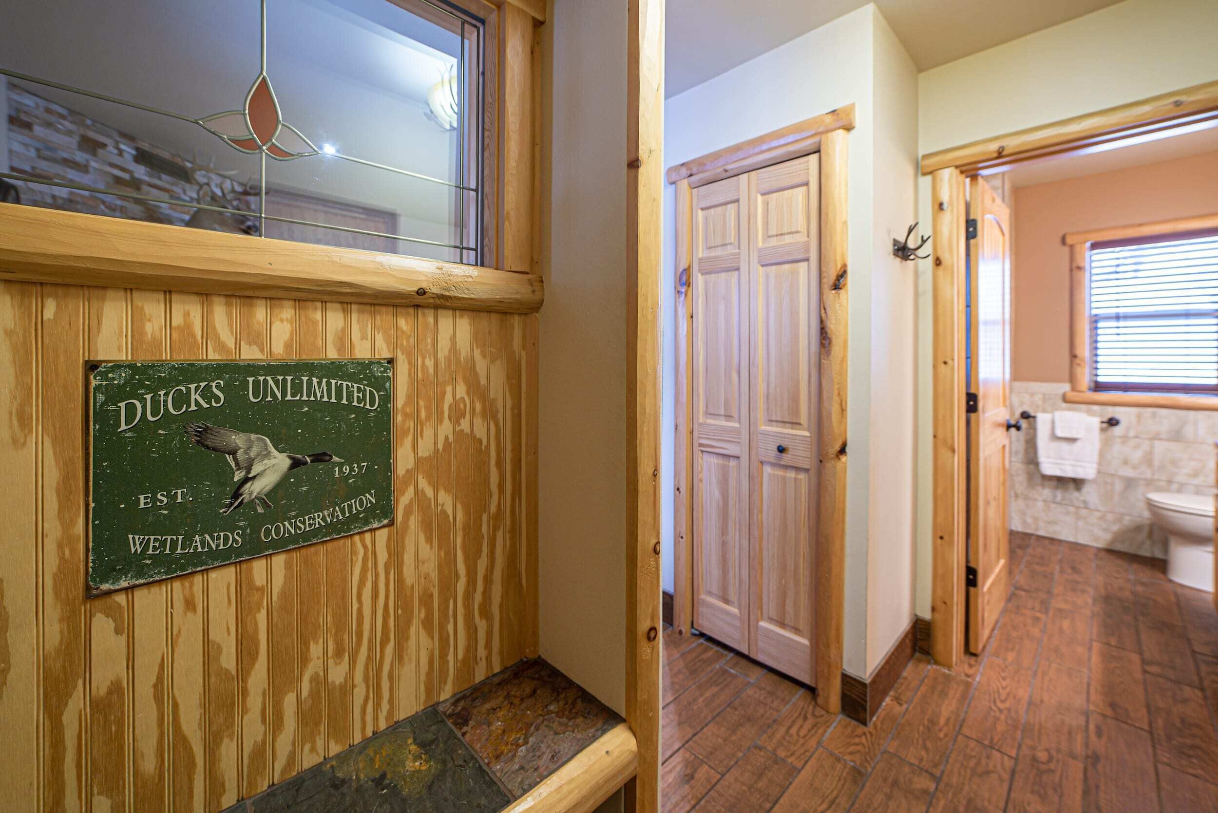 Pared de madera con un letrero que dice "DUCKS UNLIMITED EST. 1937 WETLANDS CONSERVATION" y un banco. También se muestra una puerta de madera y un baño con inodoro y ventana.