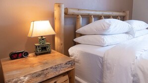 A wooden nightstand with a lamp and a digital clock, a bed with white bedding, and a wooden headboard. - The Wolverine Lodge at The Totem Lodges (Indian River)