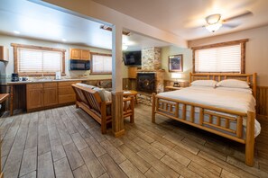 A spacious room with a wooden floor, a bed, a sofa, a fireplace, and a kitchen area with wooden cabinets and a microwave.