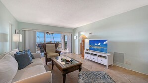 A living room with a sofa, armchair, coffee table, TV, and a view of the ocean through large windows.