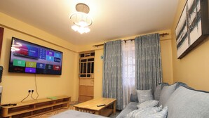 A living room with a flat-screen TV mounted on the wall, a wooden bench, a sofa, and a table. - SmartStay KAG (Naivasha)