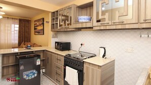 A compact kitchen with a built-in microwave, stove, and oven. There are wooden cabinets with a geometric design, and a small refrigerator. - SmartStay KAG (Naivasha)
