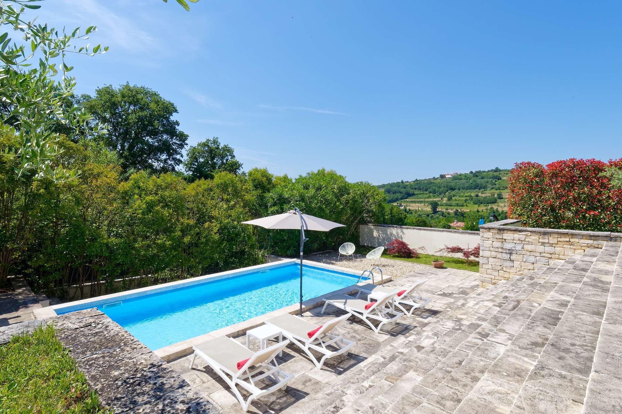 A rectangular swimming pool with clear blue water, surrounded by a stone deck with lounge chairs and a single umbrella.