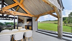 Un patio exterior con mesa y sillas de madera, una pérgola en lo alto y vistas a un campo verde.
