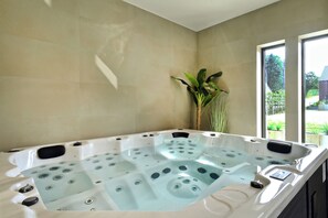 A modern spa room with a large hot tub, beige tiled walls, and a view of greenery outside. - Luxury Villa Retreat at Durbuy Suites for 12 Pers (Durbuy)