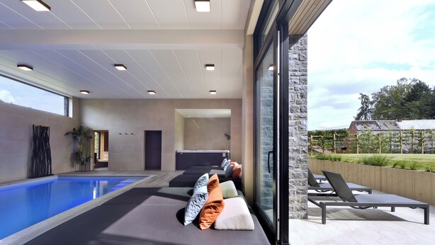 Un área de piscina interior moderna con sillones reclinables y un ventanal con vistas a un jardín.