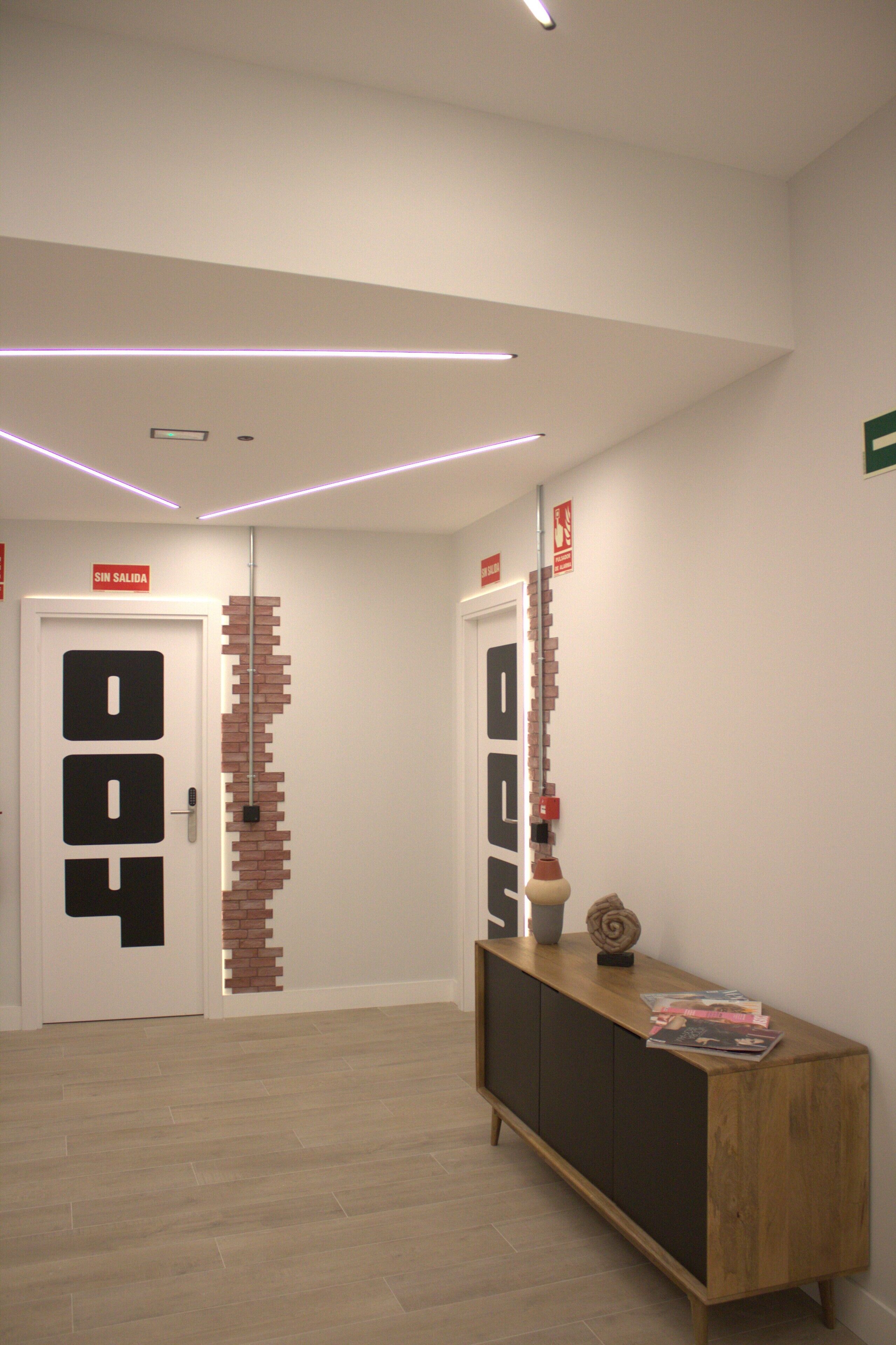 Un pasillo con una puerta blanca que indica “SIN SALIDA”, un muro de ladrillo, una consola de madera con revistas y un techo con iluminación lineal.