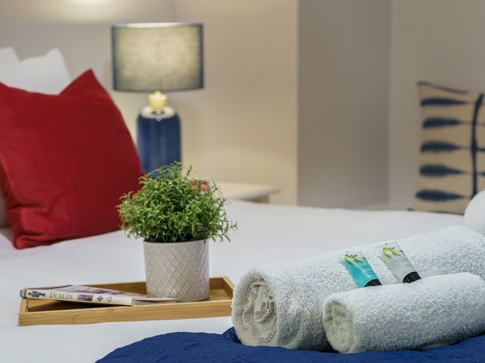 Un lit bien fait, avec une serviette blanche roulée, une petite plante en pot, un plateau avec un livre et un thermomètre, et une lampe sur une table de chevet.