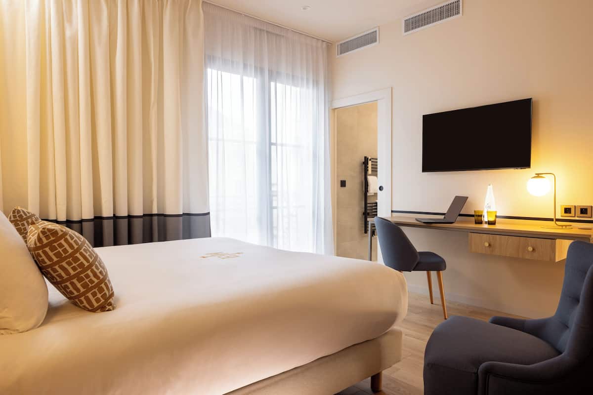 Ein Hotelzimmer mit einem Bett, einem Schreibtisch mit einem Laptop, einem Sessel und einem an der Wand befestigten Fernseher.