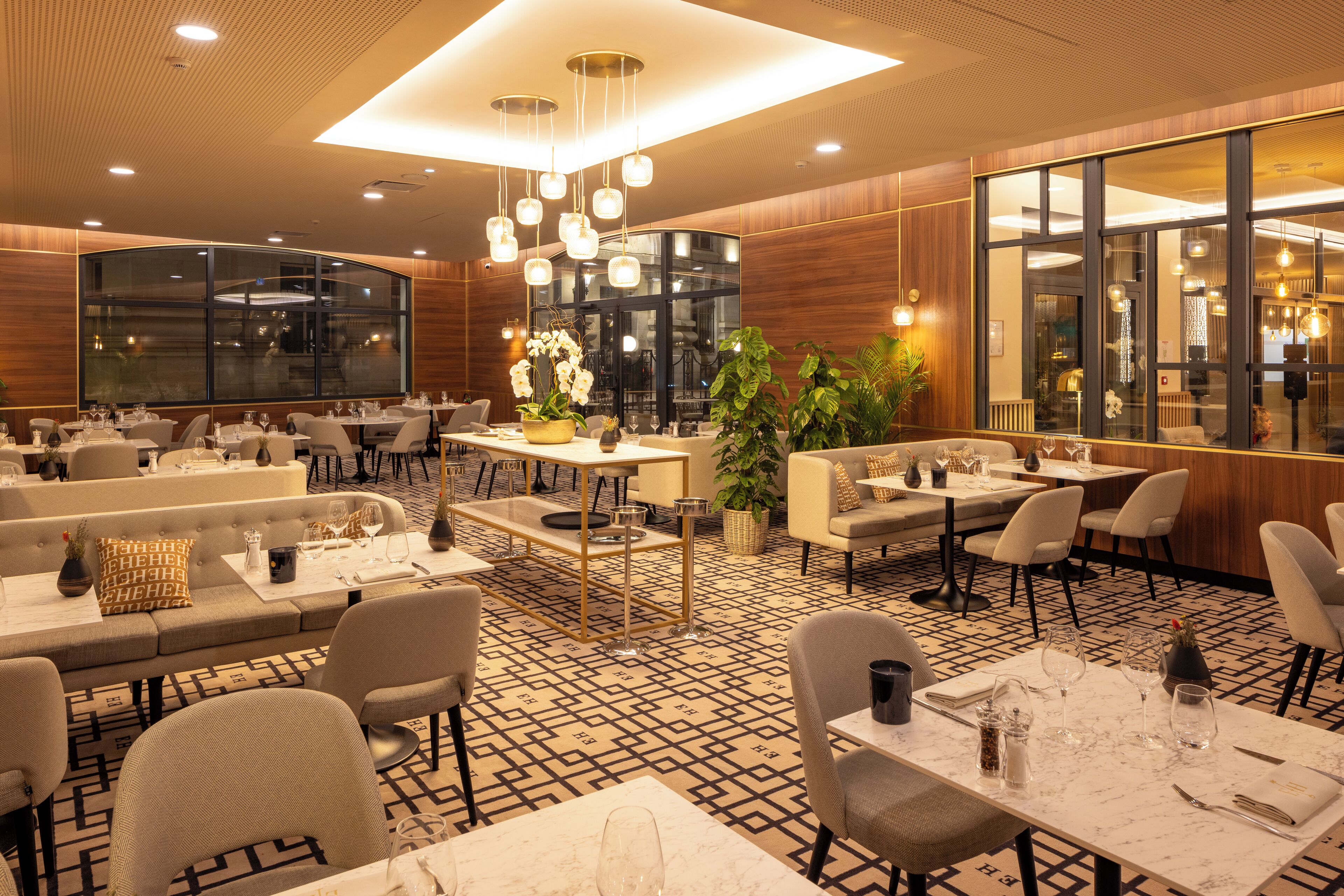 En moderne restaurant med et geometrisk mønstret gulv, flere siddeområder inklusiv borde og båse, samt et stort vindue der giver udsigt over interiøret.