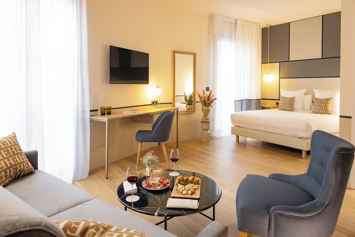 Ein modernes Hotelzimmer mit einer Couch, einem Esstisch gedeckt mit Speisen und Getränken, einem Bett, einem Fernseher und einem großen Spiegel.