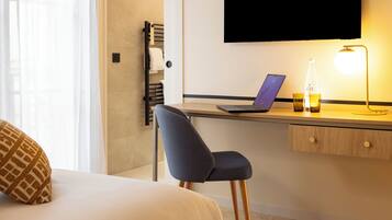 A modern hotel room with a bed, a desk with a laptop, a chair, a bottle, and a lamp.