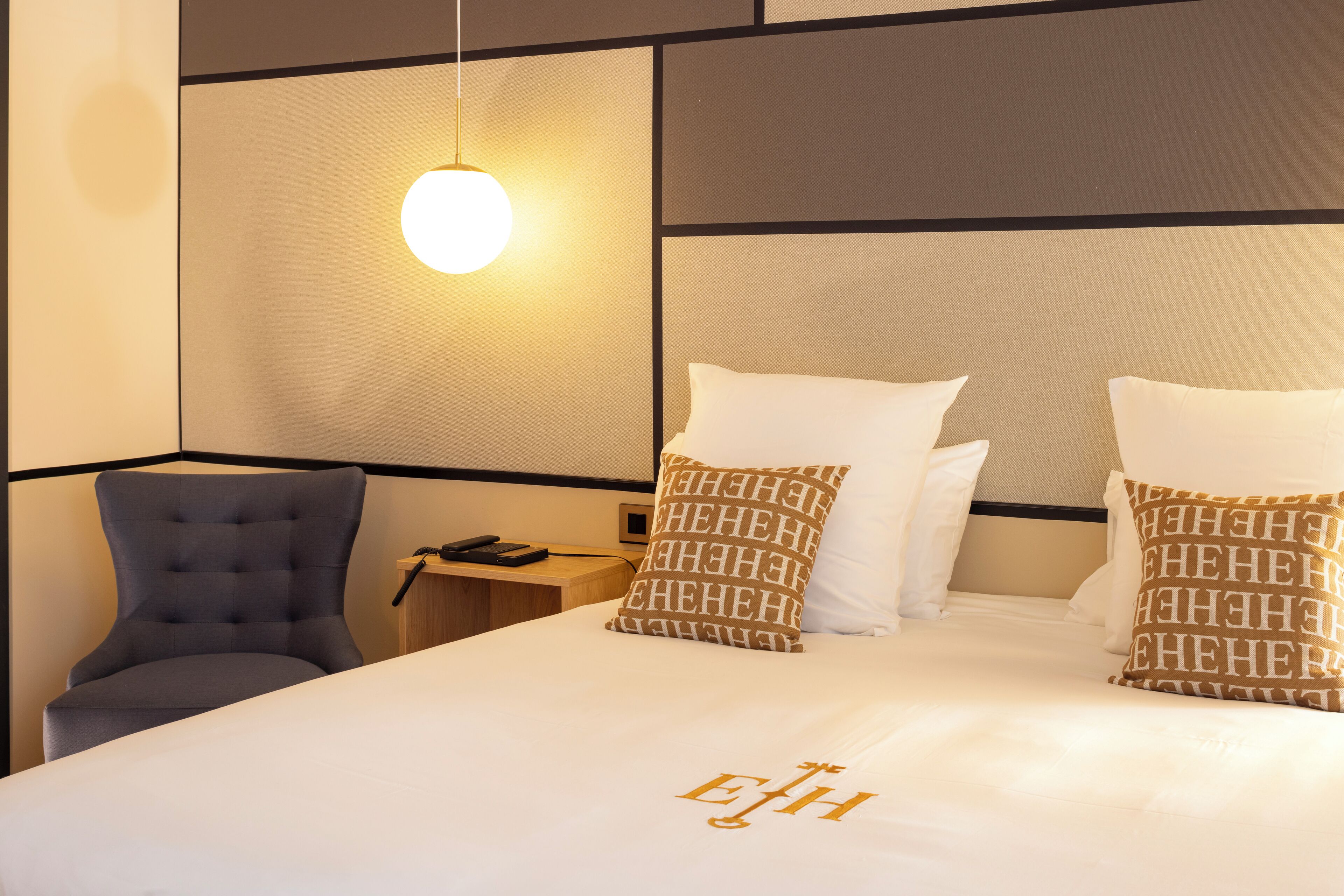 Una cama bien tendida con sábanas blancas y dos almohadas estampadas, una silla moderna y una mesita de noche con lámpara.