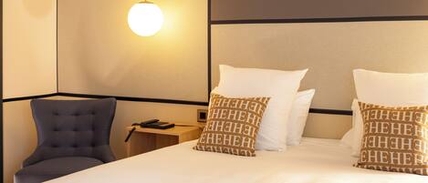 A neatly made bed with white linens and two patterned pillows, a modern chair, and a bedside table with a lamp.